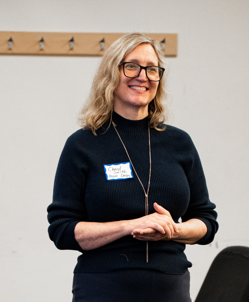 Photograph of Cheryl Smith, a white woman with shoulder length blond hair and glasses standing in a classroom.