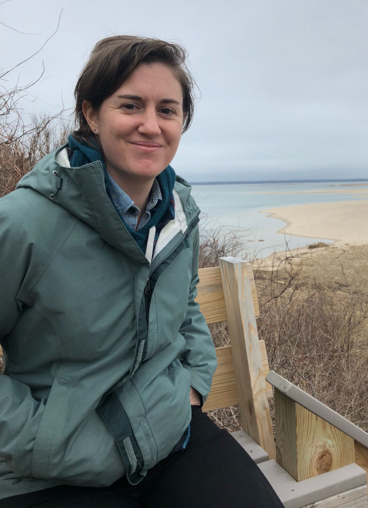 Photograph of Laurie Hurson, a white woman with short brown hair seated on a bench at the beach.